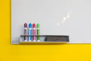 A whiteboard with markers and pens on a yellow wall.