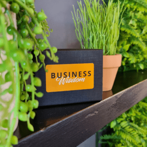 A business card from the Business Wisdom Deck by Clive Enever is sitting on a shelf next to a potted plant.