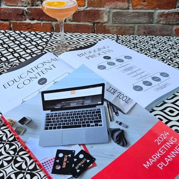 A laptop and a drink on a table next to a book, illustrating The Marketing Planner - 2024 Edition in action.