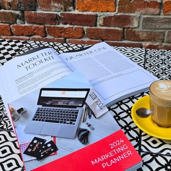 The Marketing Planner - 2024 Edition and a coffee on a table next to a laptop.