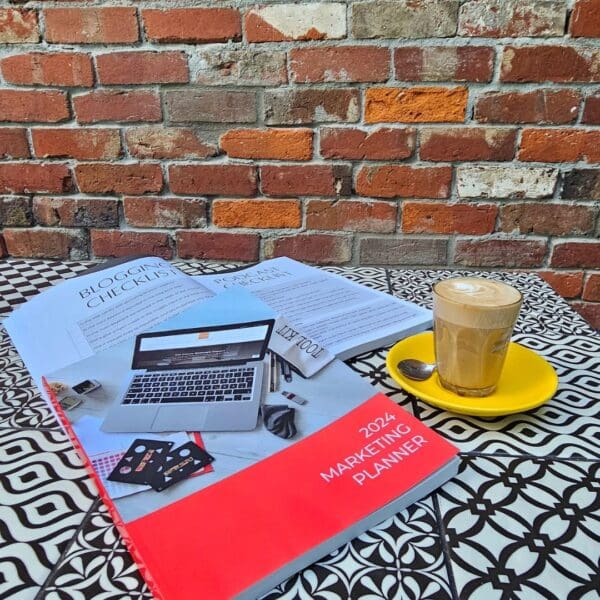 A cup of coffee and The Marketing Planner - 2024 Edition on a table in front of a brick wall, perfect for marketing planning sessions.