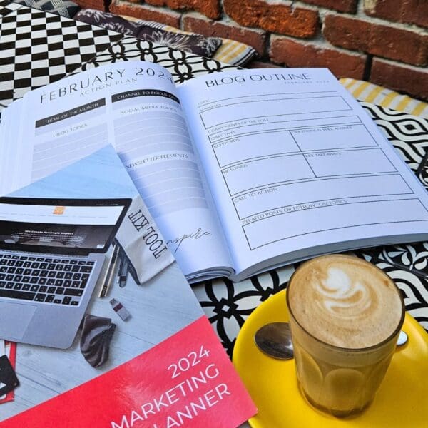 A coffee cup and The Marketing Planner - 2024 Edition book on a table next to a laptop, emphasizing marketing.